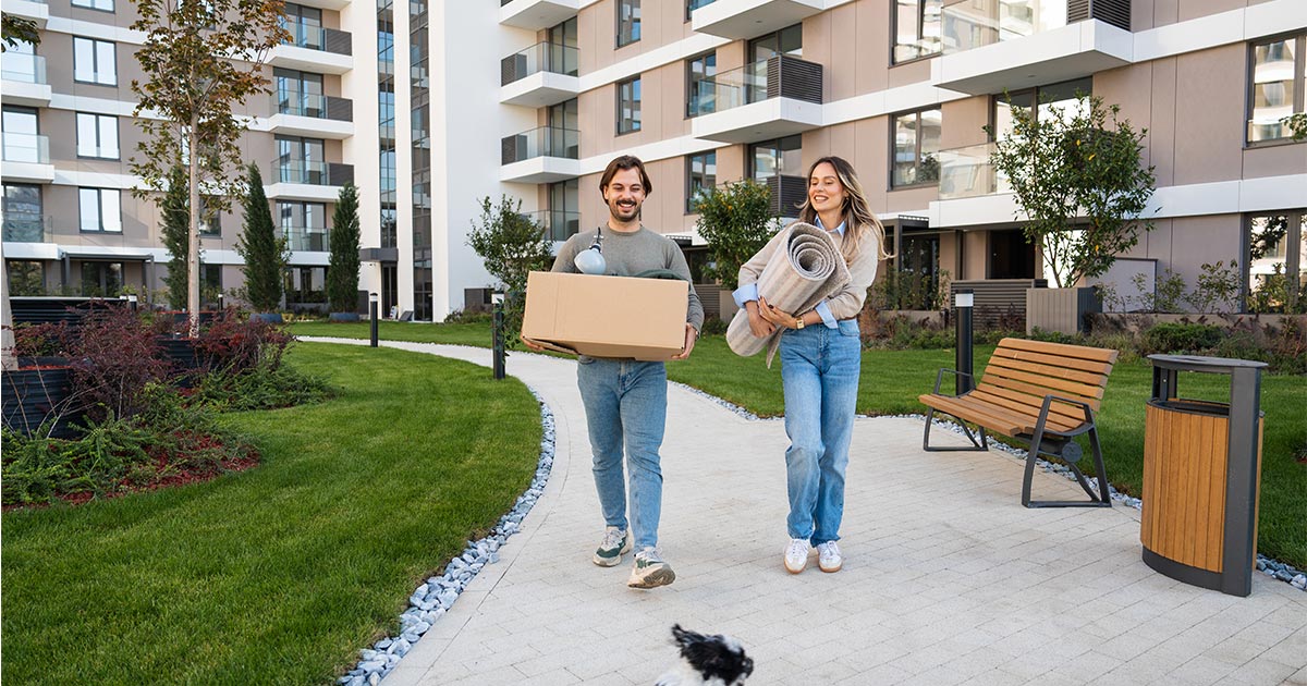 A couple walks amongst high rise apartment buildings, holding boxes, while smiling at their dog. The dog plays in the foreground. 