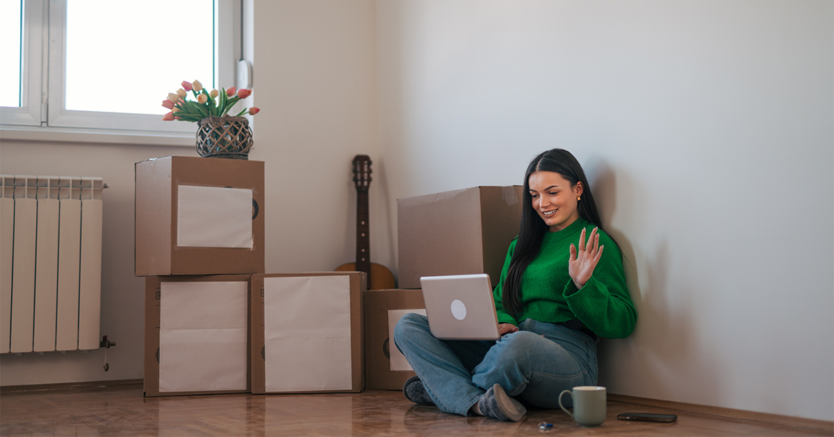 A woman, sitting next to boxes on the floor, greets a moving coordinator on a virtual call to get a long distance moving quote. 