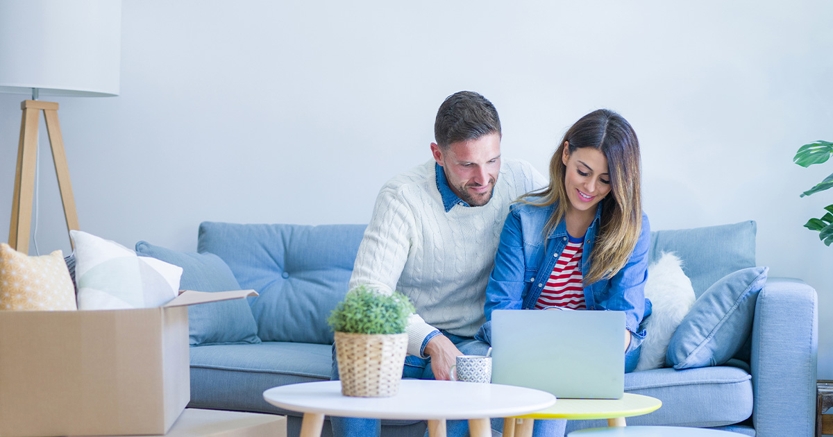 A couple, sitting on a couch, reviews their long distance moving quote options on a laptop. Moving boxes are piled near them. 