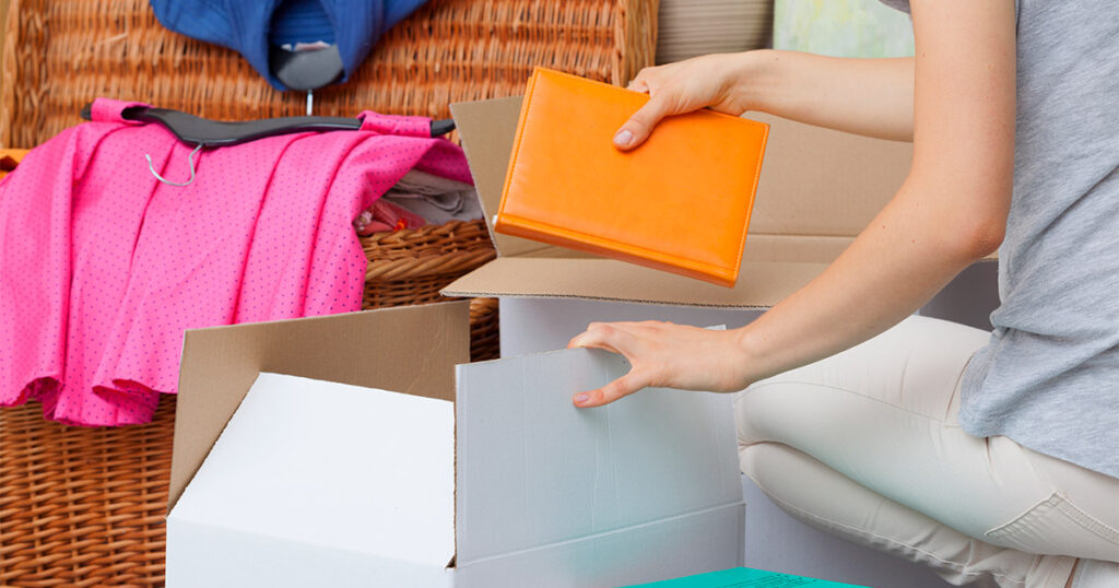 Someone sorts clothes and books into various moving boxes to declutter before a move. 