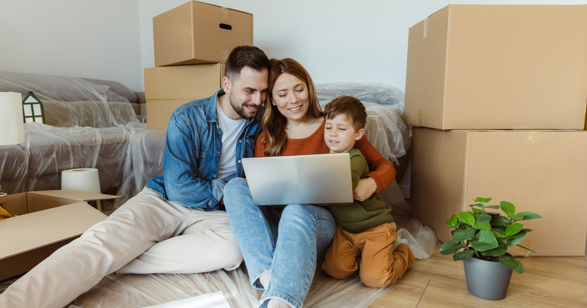 A family of three sits amongst moving boxes and packing supplies. They are looking at their new home and long distance moving company options on a laptop together.