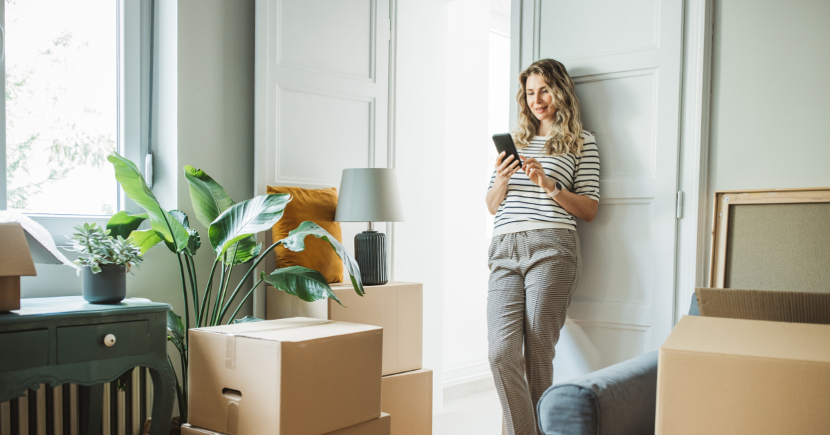 A woman, surrounded by moving boxes and packing supplies, leans against a doorway and begins to plan her move. She is using her smartphone to research whether she should use a local moving company versus a long distance moving company. 