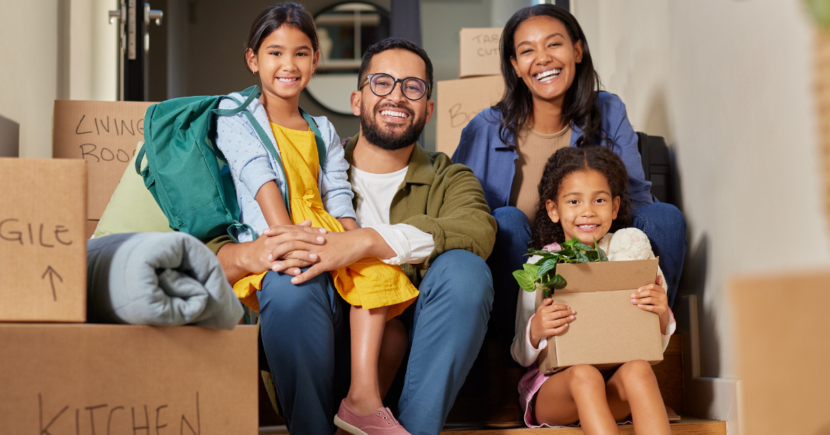 A family of four sits outside their home surrounded by moving boxes. They are going to be moving long distance to another state.