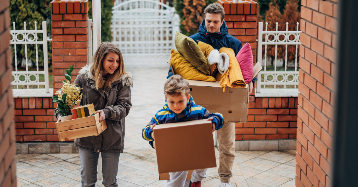 A family of three arrive at their new home after their international move. They are carrying moving boxes.