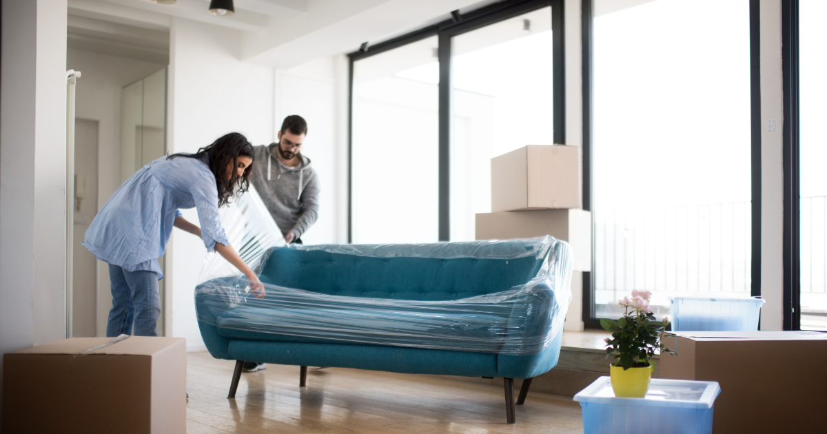 A couple begins to remove the shrink wrap on their couch in their new home after their recent long distance move. There are moving boxes surrounding them.