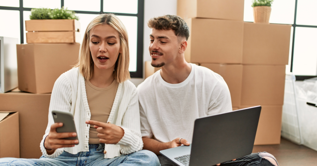 A young couple sit amongst moving boxes. They discuss long distance moving company reviews. The woman is showing a review on her smartphone, while the man is showing her a review he found on his laptop computer.