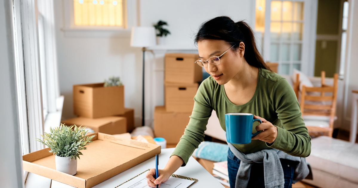 A woman holds a blue coffee mug while checking off items on her moving checklist that her long distance moving company sent to her. She is surrounded by moving boxes and packing supplies. 