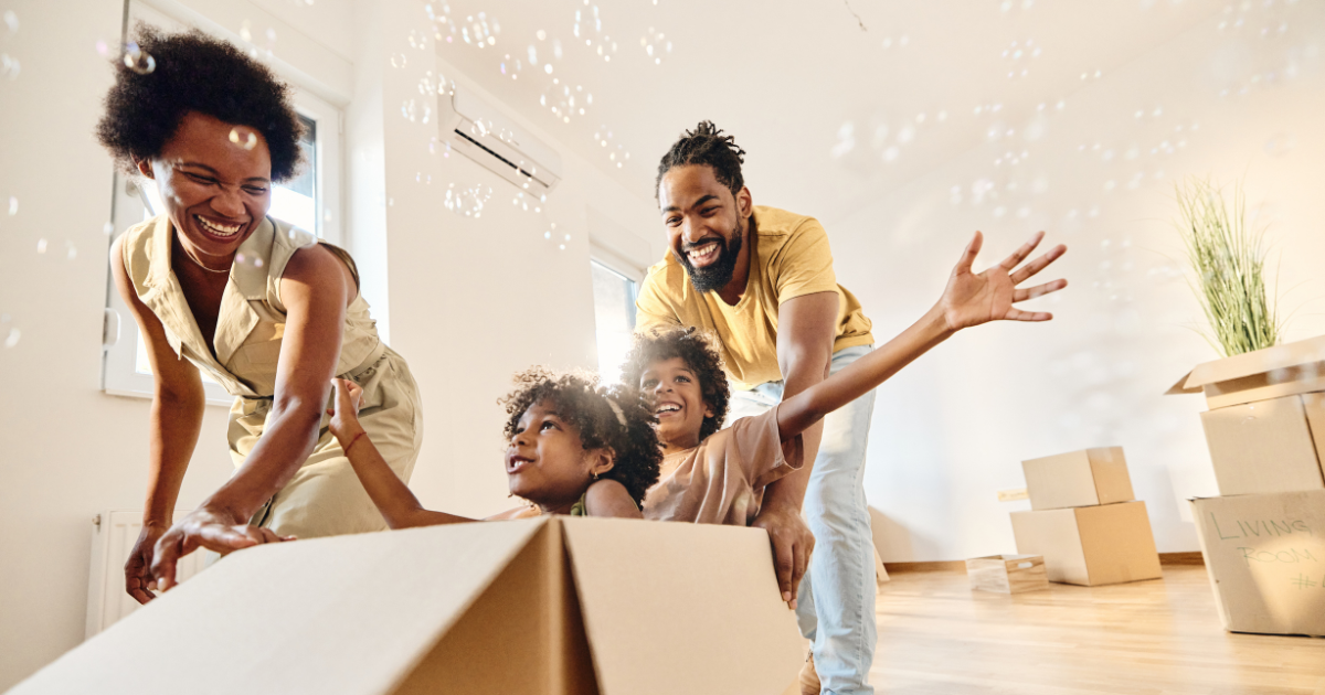 A family of four happily enjoys their new home after a long distance move. The parents play with their two children using empty moving boxes.