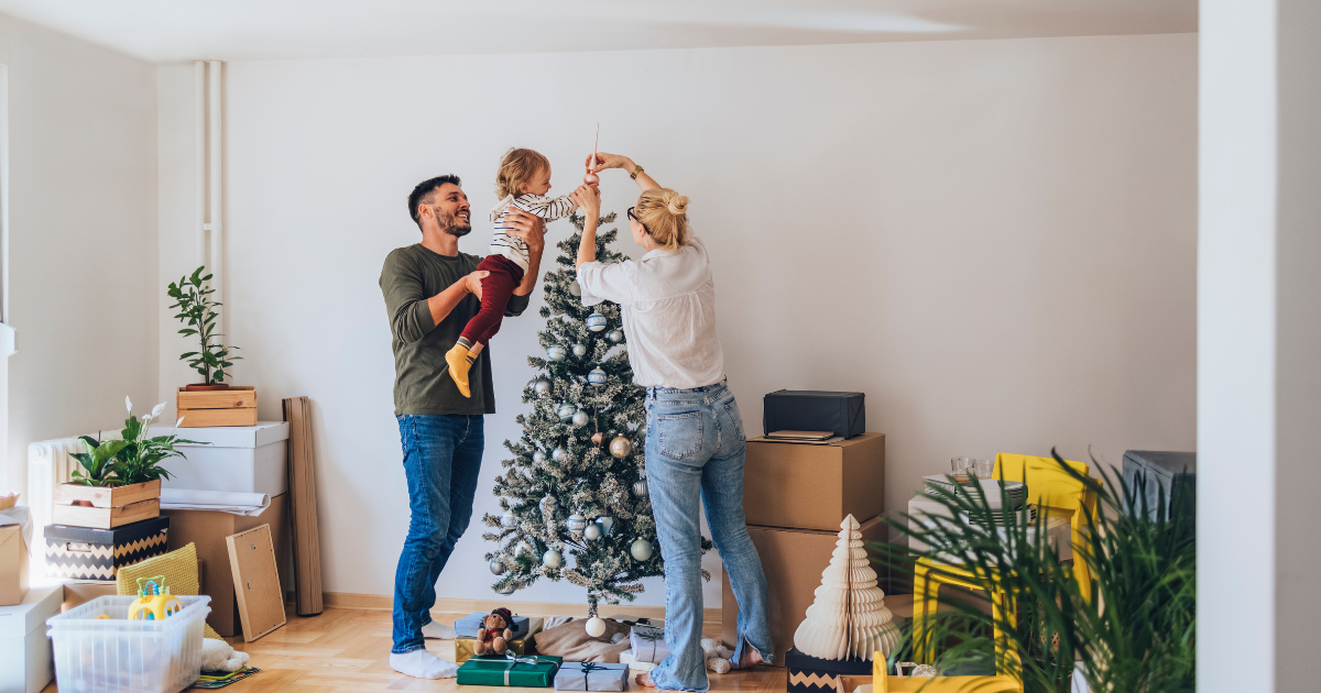 Parents decorate a Christmas tree with their young child amongst moving boxes and packing supplies.