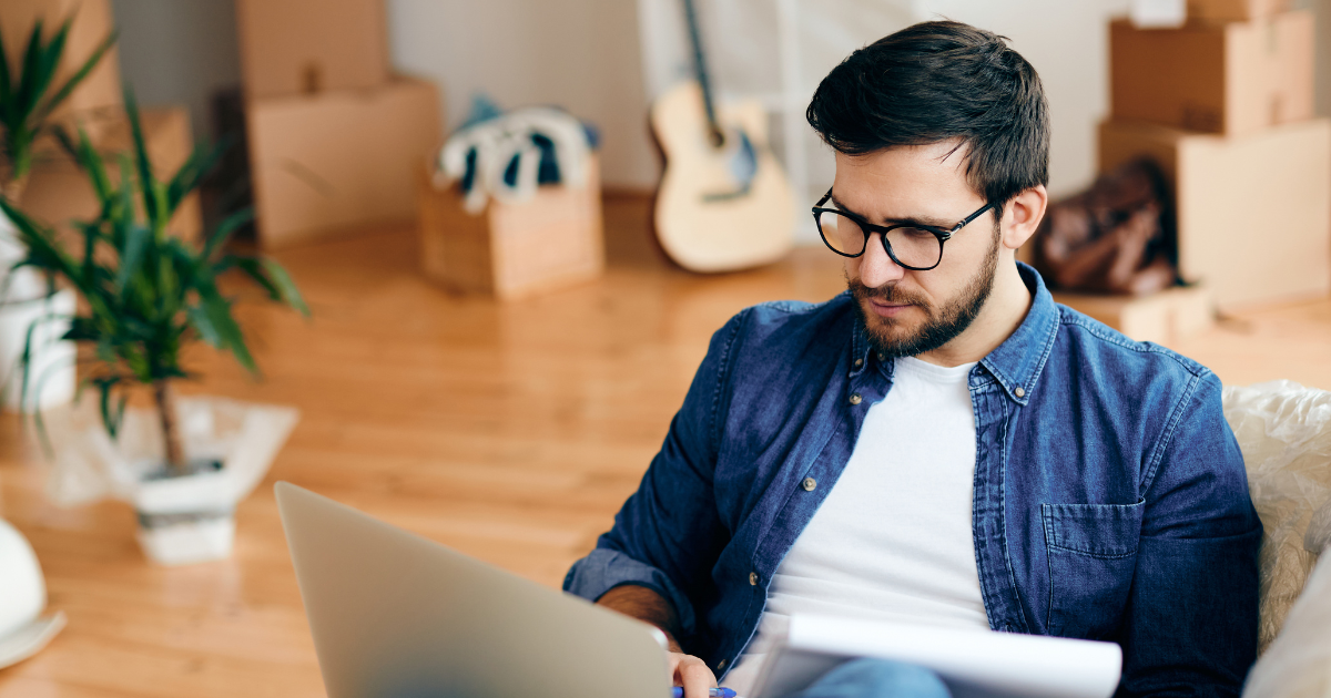 A man sits on a couch with his laptop and a notepad. He is researching BBB accredited moving companies for an upcoming long distance move. In the background, there are moving boxes and packing supplies. 