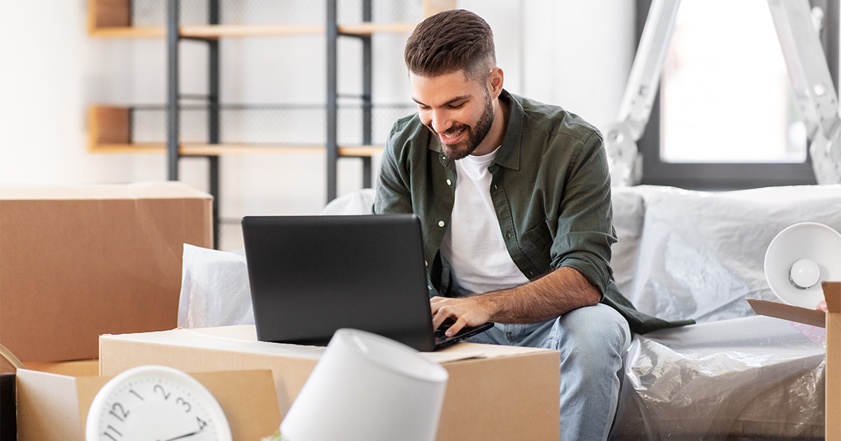 A man sits at his laptop, smiling. He is surrounded by moving boxes and packing supplies. He is actively planning his long distance move with a reputable moving company. 