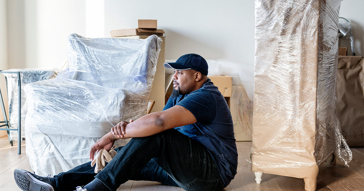 A man sits among wrapped furniture and moving boxes to take a break and focus on his breathing. He is dealing with moving stress during preparations for his upcoming long distance move. 