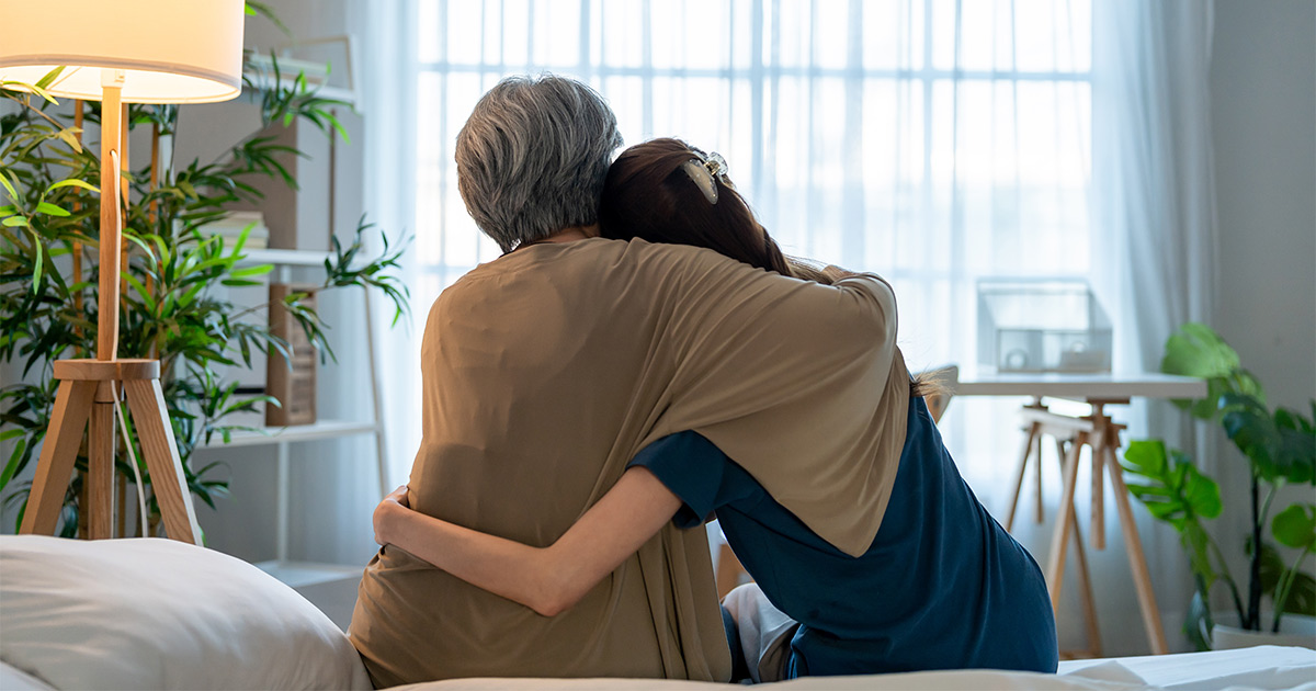 A senior and her family member sit on a bed. They are hugging. The senior is about to embark on a long distance move and is using senior movers to help out. 