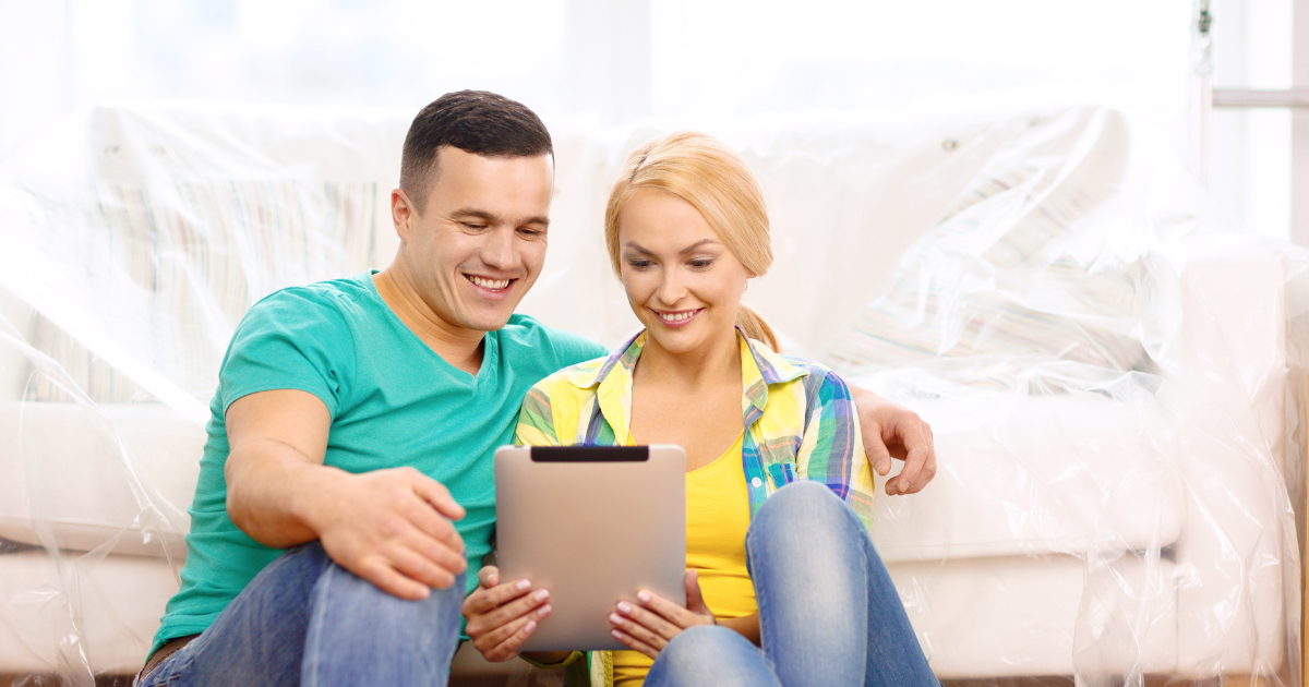 A man and woman sit together in front of a couch wrapped in plastic. They are reviewing moving quotes from affordable moving companies for their upcoming long distance move on a tablet. They are smiling.