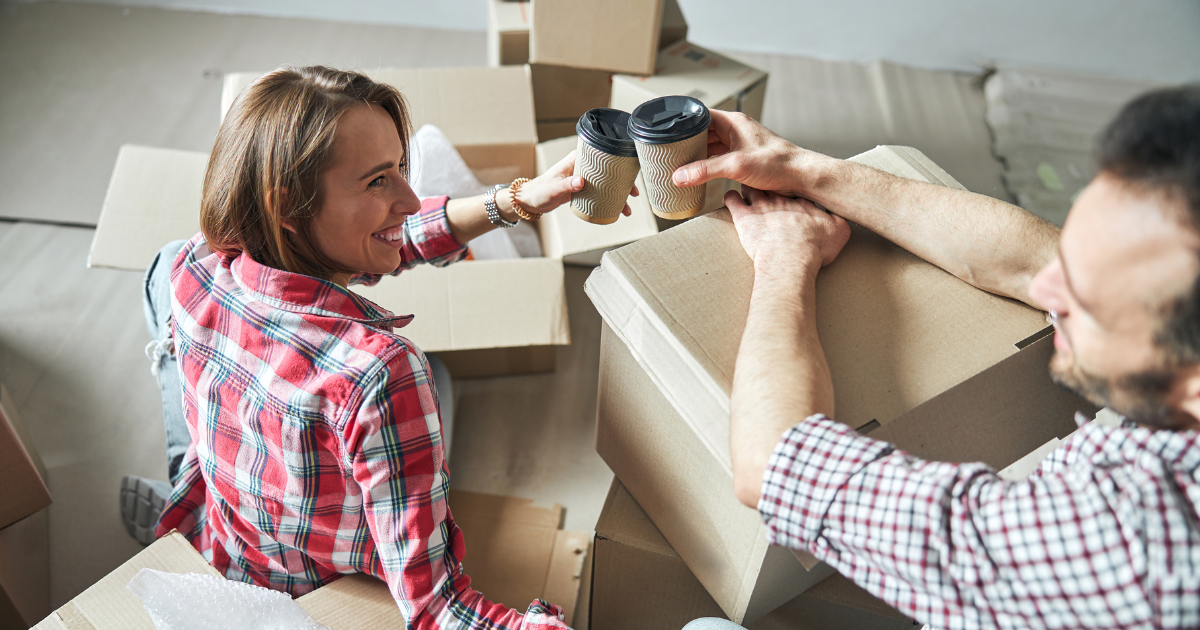 A man and woman sit amongst moving boxes and packing supplies. They are raising a toast with their coffee cups to packing their own stuff for a long distance move. They are smiling.