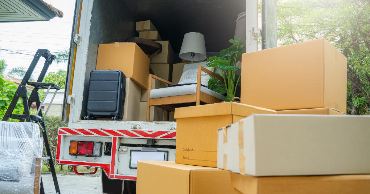 The back of a moving truck. The door is open, and inside are some pieces of furniture and moving boxes. There are more moving boxes that sit on the pavement next to the moving truck. This is occurring after a long distance move.