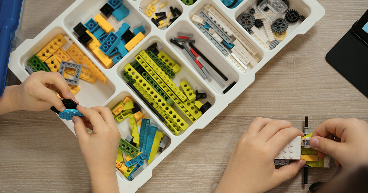 Two children disassemble and sort their Lego bricks into a storage bin by size and color. They are preparing for a long distance move.