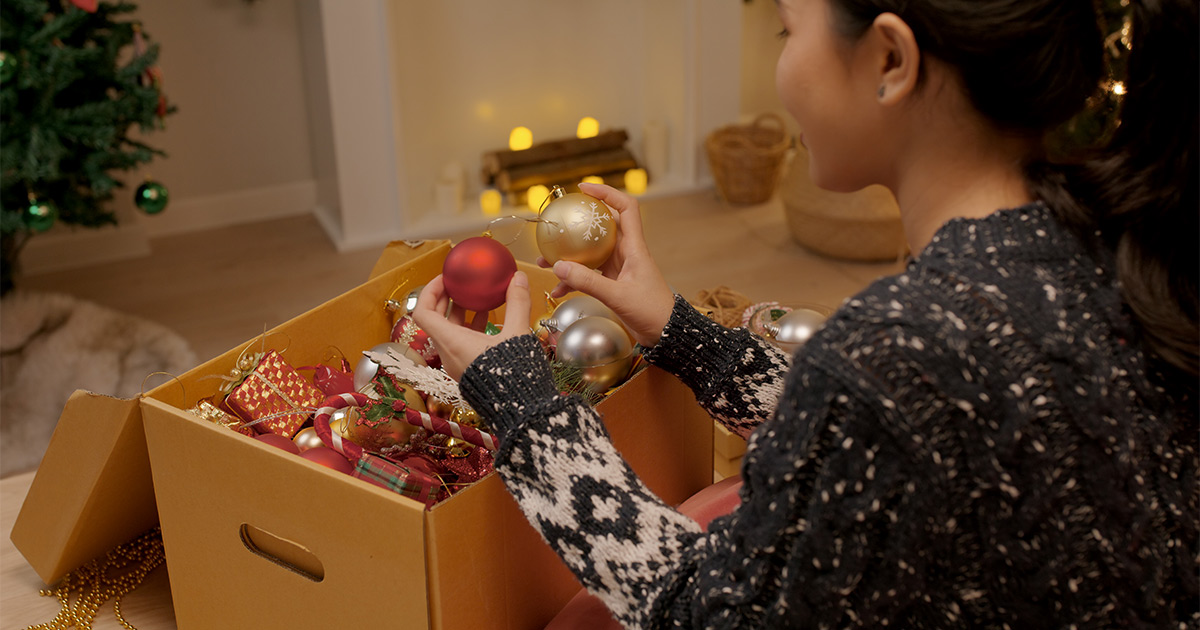 A young woman in a sweater looks at ornaments in a box next to a Christmas tree. She is using Christmas decor storage to keep everything organized. She will be moving cross country soon. 