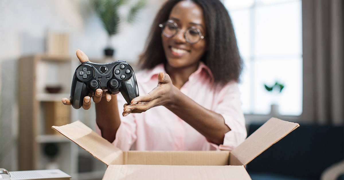 A woman sits in front of an open moving box while smiling. She is putting a video gaming controller into the box for her upcoming move. She already knows how to pack a PC for moving and is making sure her gaming collection arrives in tact.