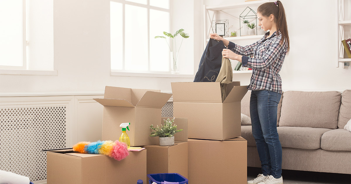 A young woman unpacks moving boxes in her new living room after an emergency move. There are also cleaning supplies around and on top of the boxes.