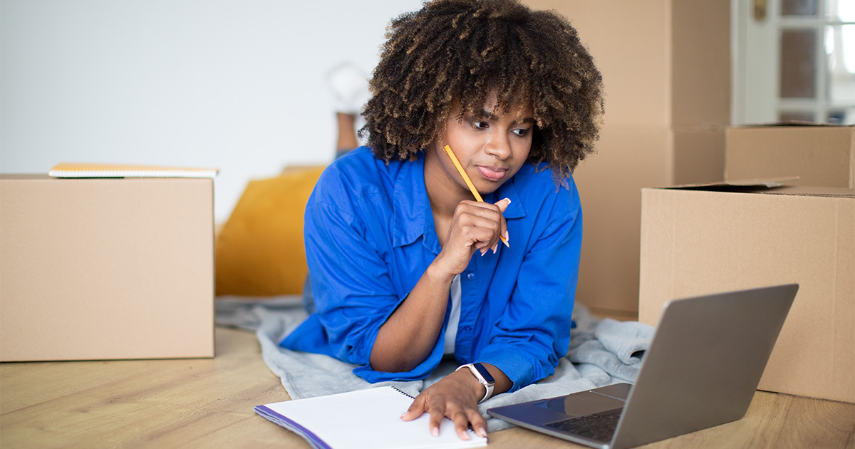 A young woman lies on the floor with her laptop amongst moving boxes. She is holding a pencil and has some paper next to her. She is contemplating her options for moving on a budget for her upcoming cross country move.