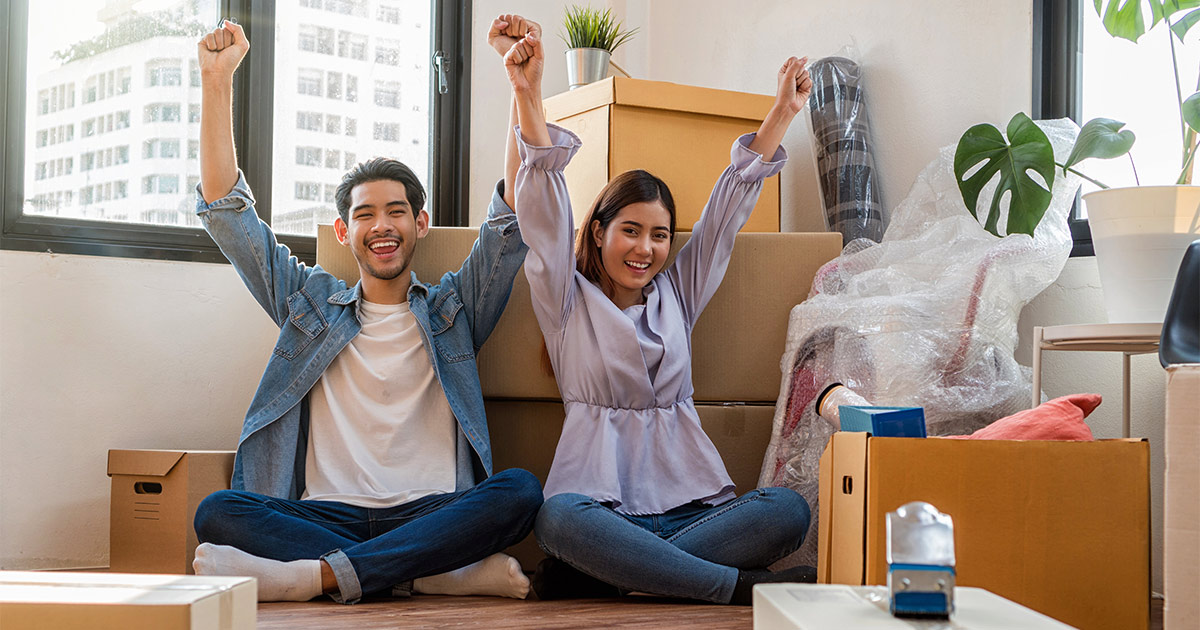 A young couple sit on the floor of their new apartment after a cross country move. They are surrounded by moving boxes and packing supplies. They are raising their arms and smiling. They figured out the cheapest way to move cross country through careful research and planning.