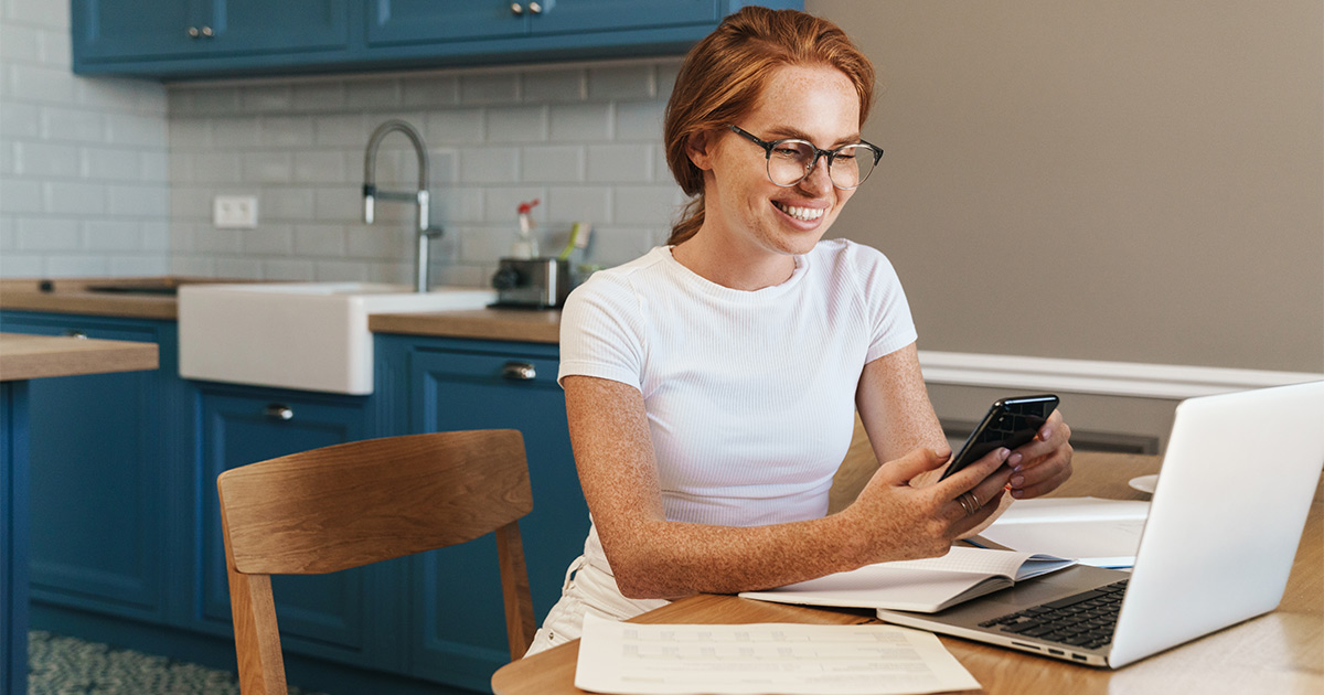 A woman sits at her kitchen table with her smartphone and laptop, with papers and a notebook in front of her. She is smiling while she looks at her phone. From her research online, she knows it will be more beneficial to use a full-service moving company for her small move.