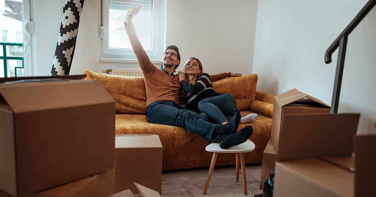 A happy couple sits on their couch in their new home after a cross country move. They are surrounded by some moving boxes. They used a full-service moving company for convenience.
