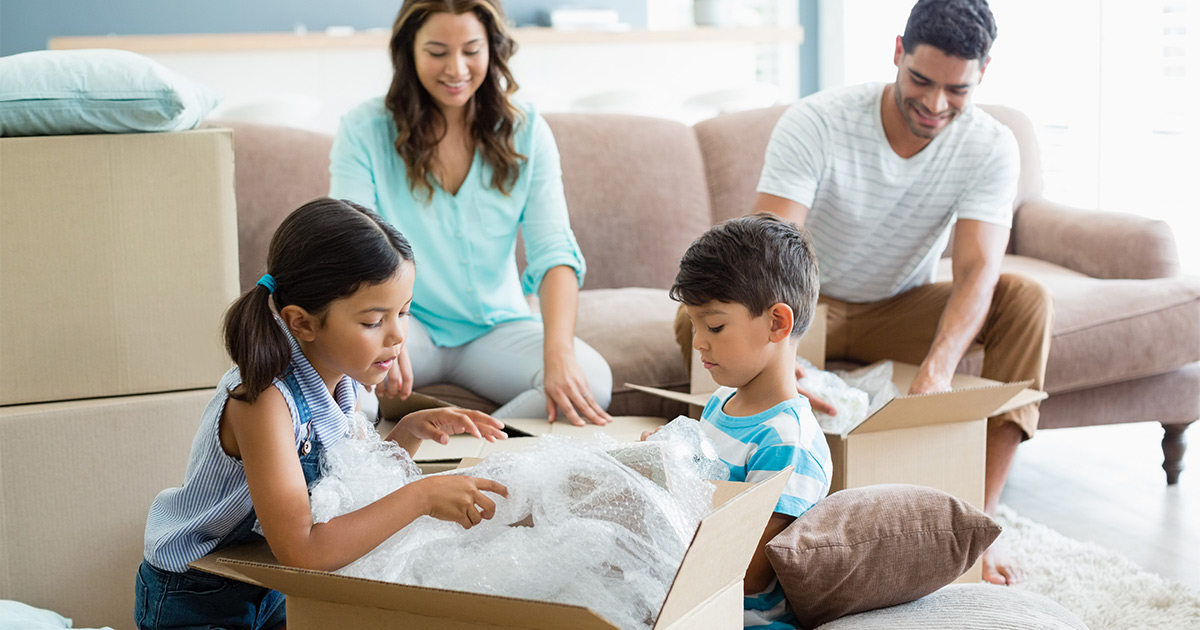 A family of four are packing items in moving boxes in their living room. They are preparing to use a full-service mover for their upcoming move.