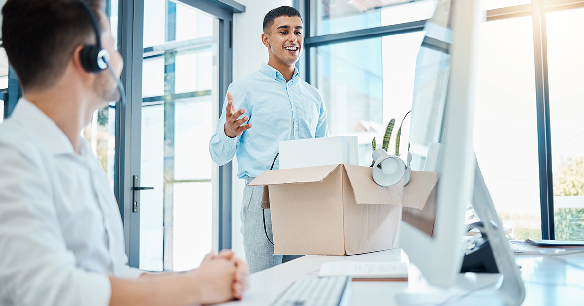 A man smiles while talking to a coworker, who is at his desk working on a computer. A box of office supplies and decorations sit on the desk in front of the smiling man. This is his first day at his new job; he has relocated for work.