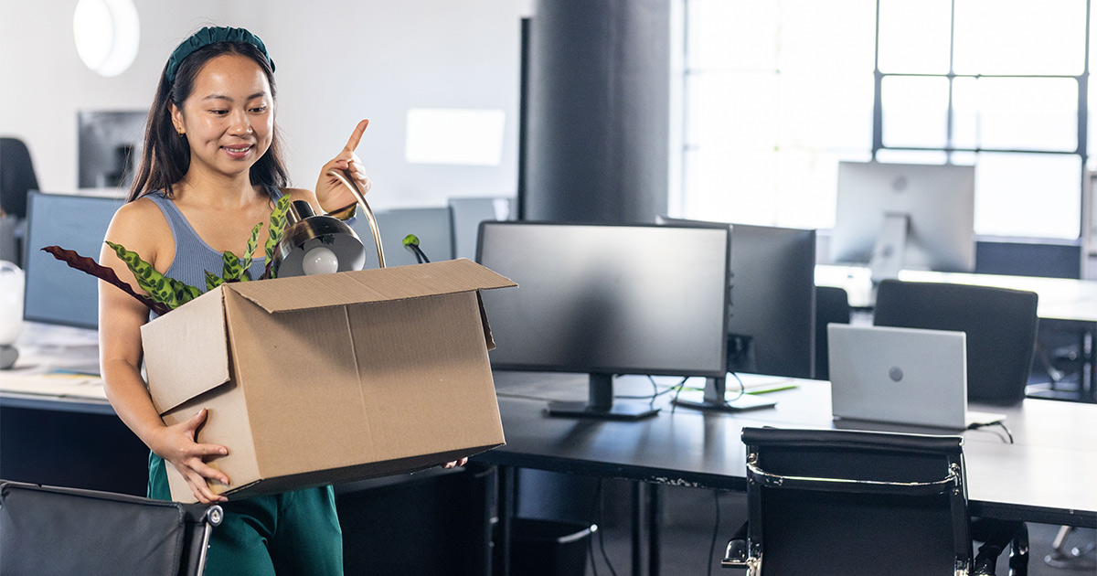 A woman carries a moving box full of office supplies through an office. She has just accepted a new job and will be relocating for work, so she's clearing her desk off.