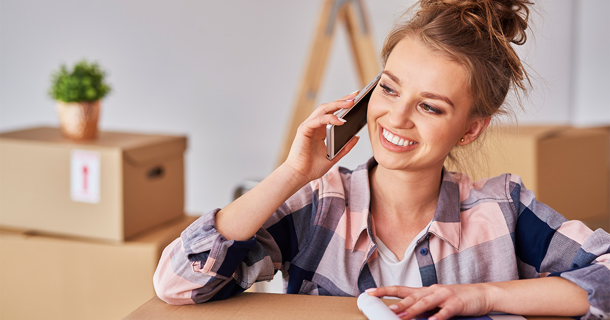 A young woman is smiling while taking a call on her smartphone. She is sitting amongst moving boxes. She is talking to her real estate agent about the new details of her closing date. 