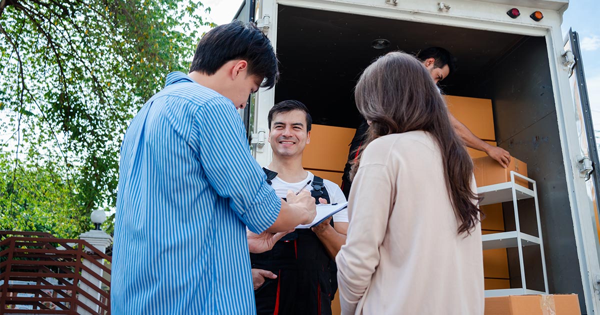 A married couple signs paperwork with a mover in overalls. They are near an open moving truck. The couple is moving out of the country to Europe. They are using an international mover. 