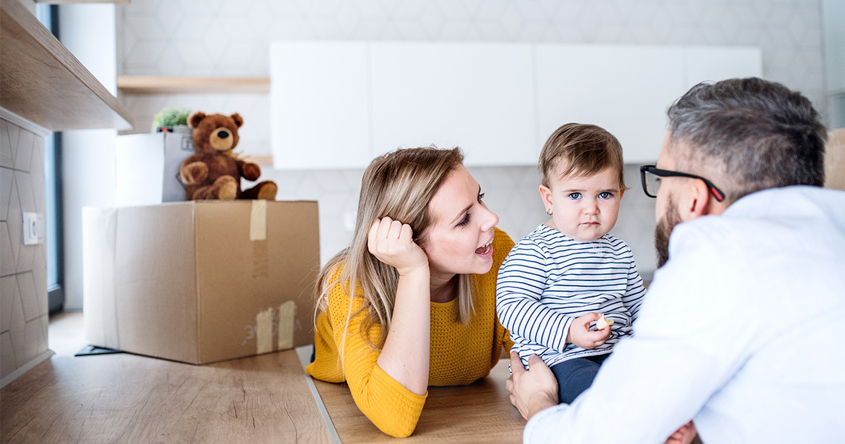 Parents play with their infant child in the kitchen. A moving box sits on the counter with a teddy bear on top. They are preparing for a cross country move, and are using a partial-service moving company.