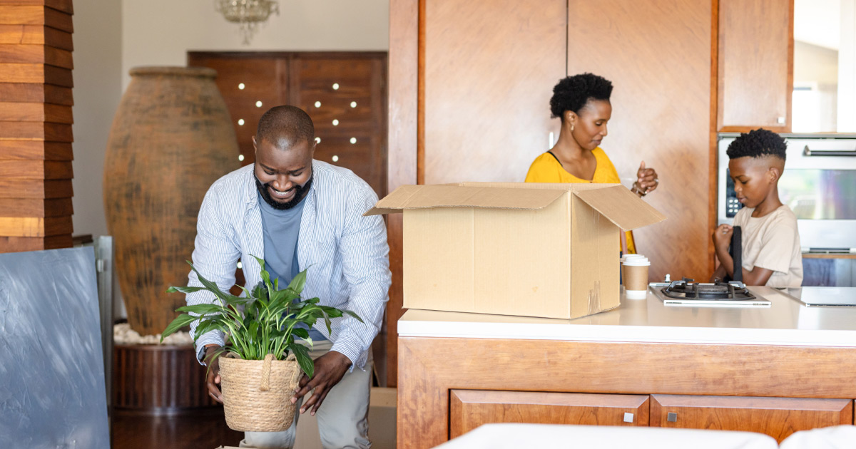 A family of three begins to pack belongings in their home for a cross country move. They are in the kitchen, and the father smiles while arranging a plant for moving. They are using partial-service movers. 