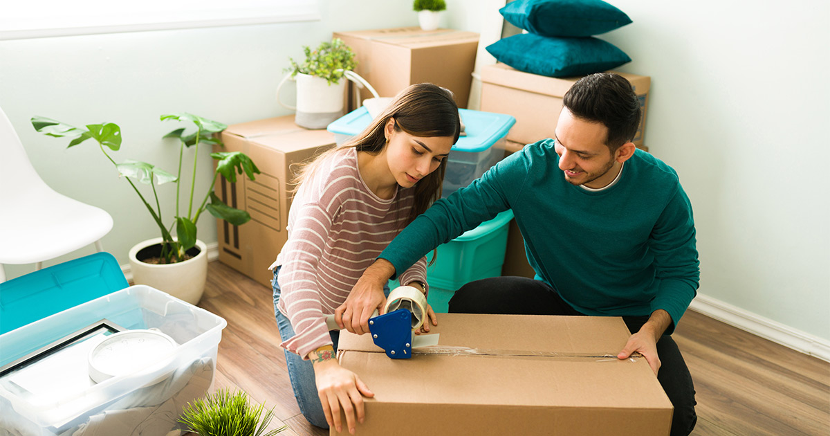 A young couple, surrounded by moving boxes and packing supplies, tapes up a box. They are doing a self pack option with their moving company for an upcoming cross country move. 