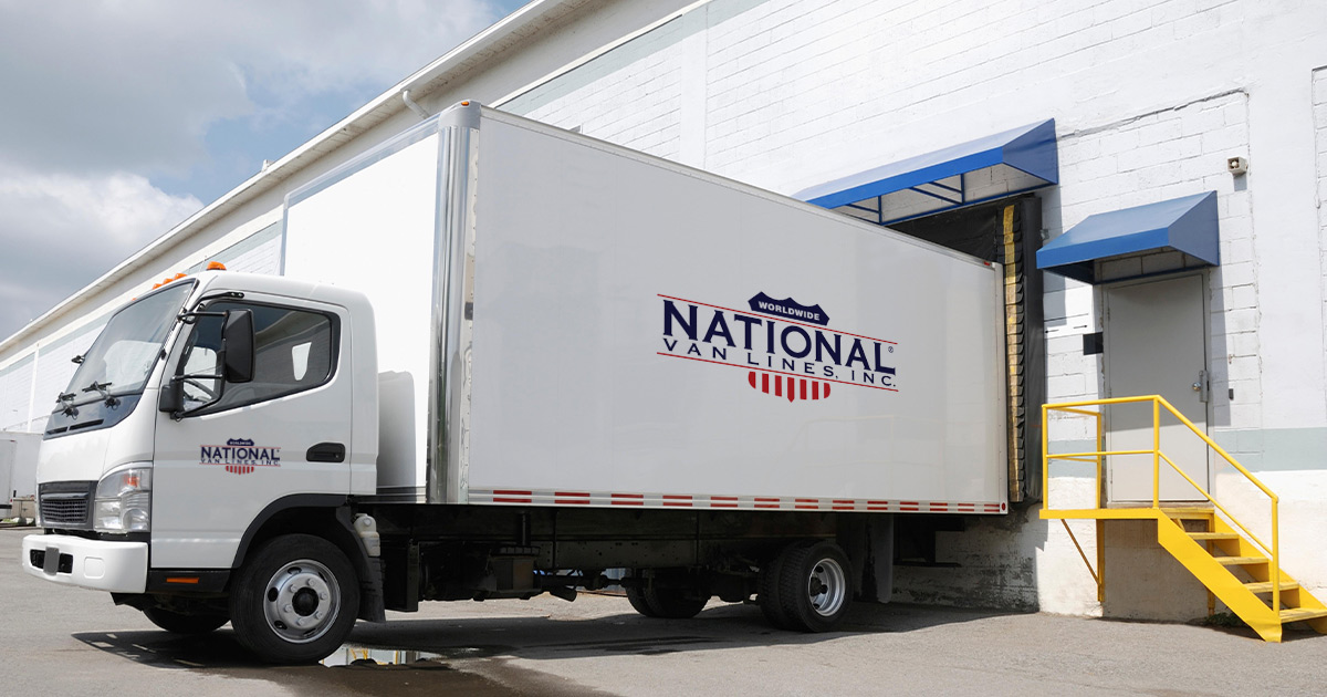 A National Van Lines truck is shown backed up to a storage facility. It is dropping off a load of boxes that are being stored during a long distance move.