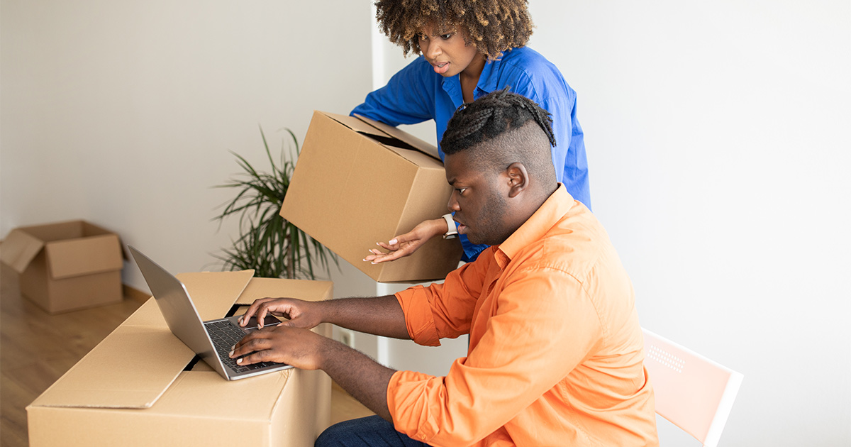 A couple has a laptop on top of a moving box. The man sits at a large box with a chair and uses it as a surface to use his laptop. The woman holds a moving box and looks at the screen with him. They are using Reddit moving tips to help determine what long distance moving company they want to use.