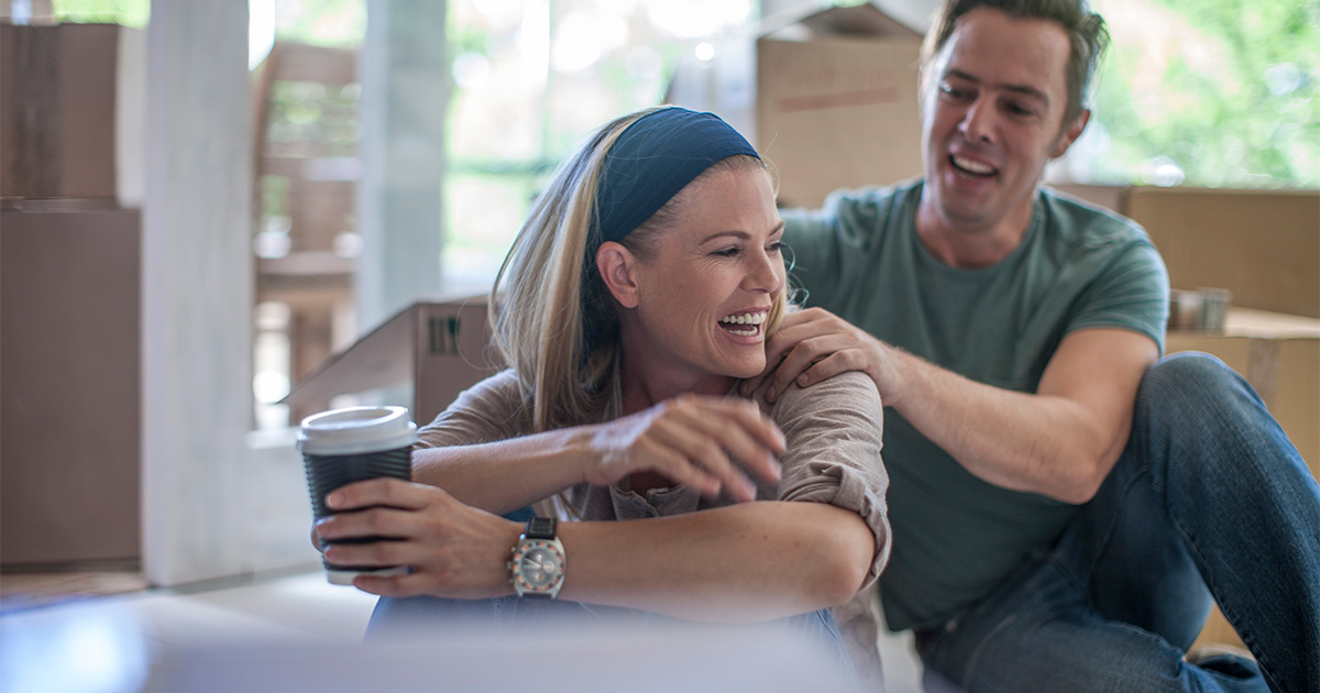 A couple sits amongst moving boxes in their living room. They are preparing to move long distance. They are smiling and supporting each other. The woman holds a cup of coffee. They used Reddit moving tips to help determine the best options for moving companies.