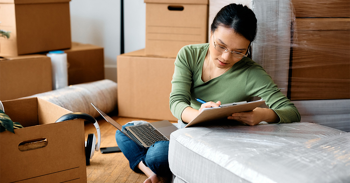 A woman sits amongst moving boxes and packing supplies on the floor. She has a checklist and a laptop out. She is moving coast to coast and is ensuring she stays organized along the way. 