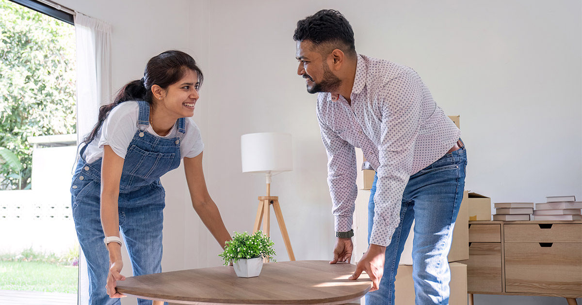 A couple smiles at each other while placing a coffee table in the living room of their new home. There are some moving boxes behind them. They had a great experience with their moving company, because they learned how to use Yelp to review moving companies. 