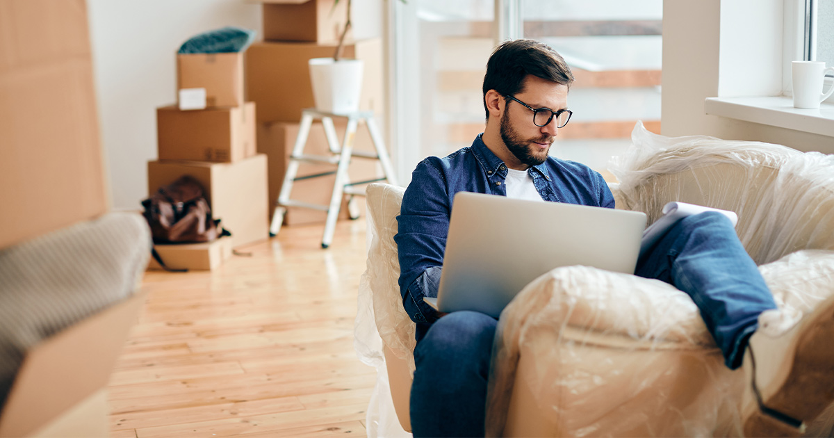 A man sits on his plastic wrapped couch. Moving boxes and packing supplies are in the background. He is reviewing a notepad and has a laptop. He is doing research with tools like Yelp to determine a good moving company for his long distance move. 