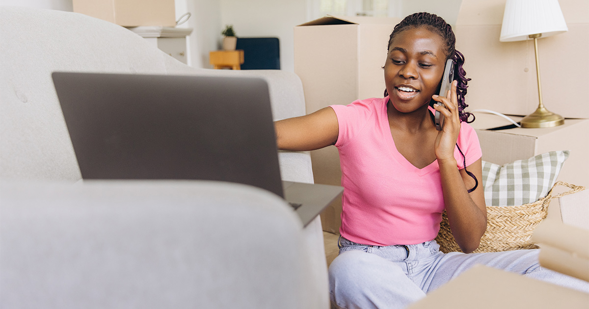 A woman sits in her living room amongst moving boxes on the floor. Her laptop is set up in front of her on her couch, and she is on the phone speaking with a moving company. She used websites like Yelp to find moving reviews and select a moving company. 